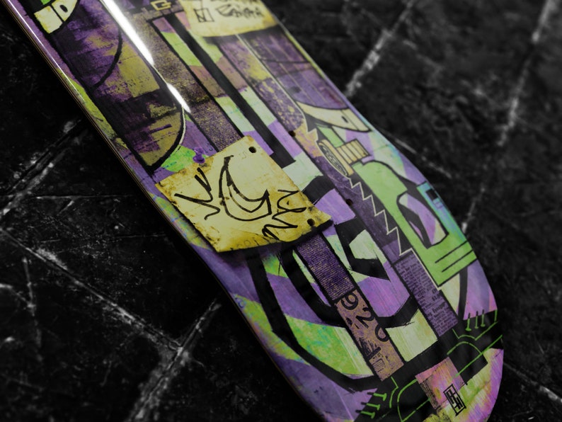 May include: A purple, green, and yellow skateboard deck with a graphic design featuring abstract shapes and a small yellow sticker with a crescent moon and a bird design.