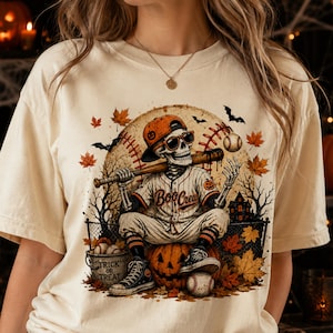 May include: A cream-coloured t-shirt featuring a graphic of a skeleton wearing a baseball cap and sunglasses, holding a bat. The skeleton sits on a pumpkin, with text reading "Boo Crew" and "Trick or Treat" on a bucket, surrounded by autumn leaves.