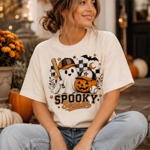 May include: A cream-colored short-sleeve t-shirt featuring a graphic print of a ghost and a jack-o'-lantern wearing baseball caps. The design includes a baseball bat, a baseball, a bat, stars, and the text "SPOOKY Season" in orange and black.