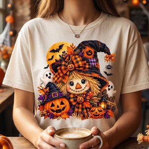 Può includere: Una t-shirt color crema con la grafica di uno spaventapasseri che indossa un grande cappello da strega scuro a quadri, decorato con fiocchi arancioni. Il disegno include due zucche di Halloween, due fantasmi bianchi, una luna gialla, pipistrelli.