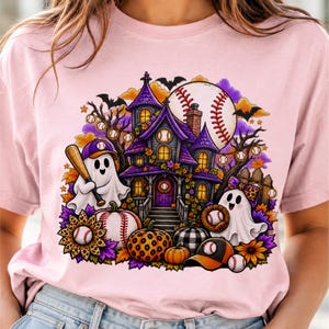 May include: A light pink t-shirt featuring a graphic of a purple haunted house, two ghosts, pumpkins, and baseball elements. One ghost holds a baseball bat, while the other wears a leopard print bow. The design includes baseballs, bats, and bats.