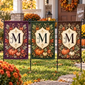Puede incluir: Tres banderas de jardín rectangulares con un escudo central color crema con la letra M en fuente serif. Cada bandera está rodeada de hojas de otoño, bellotas y calabazas sobre fondos de color morado, verde y marrón.
