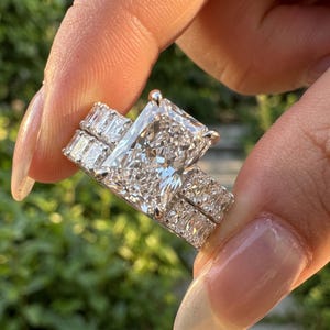 May include: A silver-toned ring featuring a large rectangular radiant-cut center stone held by four prongs, flanked on each side by a row of three smaller rectangular baguette-cut stones set into the band, creating a multi-stone design.