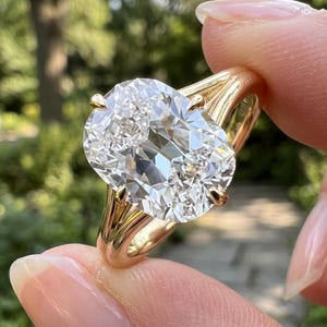 May include: A gold ring featuring a large, clear, cushion-cut gemstone held by four prongs. The band is smooth and polished, with a split-shank design that connects to the setting holding the central stone securely in place.