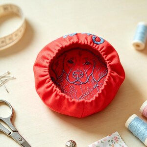 May include: A red fabric pincushion featuring an embroidered golden retriever face, accompanied by a metal thimble, small silver scissors, a measuring tape, several sewing pins, spools of thread in various colours, and a small floral fabric square.