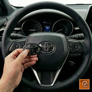 May include: A black Toyota emblem overlay held in front of a chrome Toyota steering wheel logo. The steering wheel is black leather with integrated control buttons and a digital instrument cluster visible in the background of the vehicle interior.