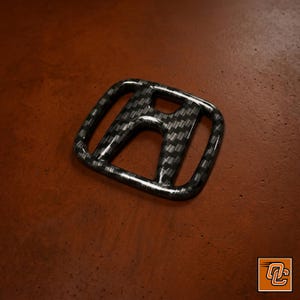 May include: A Honda emblem badge featuring a glossy black and grey carbon fibre weave pattern. The iconic H-shaped logo has a smooth, reflective surface with a distinct checkered texture throughout the entire frame of the automotive insignia.