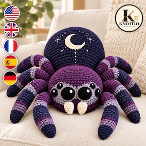 Puede incluir: Una araña de ganchillo con un abdomen morado oscuro bordado con una luna creciente y estrellas blancas. La cabeza es de color morado claro con ocho ojos negros y dos colmillos color crema. Las ocho patas tienen rayas moradas, lavanda y azul oscuro.