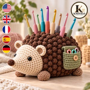 Könnte beinhalten: Ein braun-cremefarbener Häkel-Igel-Organizer mit einem strukturierten Rücken, der neun bunte Metall-Häkelnadeln hält. An der Seite befindet sich eine kleine grüne Tasche mit einem Holzknopf, die drei kleine Metall-Maschenmarkierer enthält.