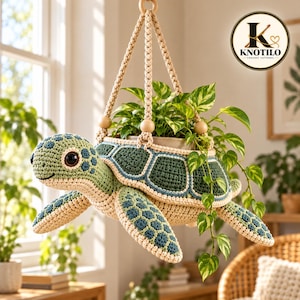 Può includere: Un portavasi sospeso all'uncinetto a forma di tartaruga marina con testa verde chiaro, parte inferiore color crema e guscio verde scuro con motivi esagonali bordati di blu. La tartaruga contiene una pianta in vaso con foglie verdi.