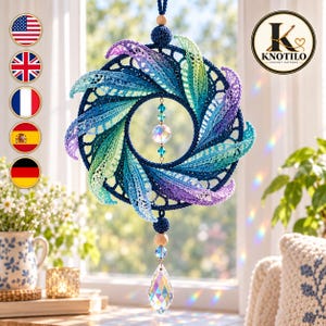 May include: A circular crochet wall hanging featuring a dark blue ring adorned with radiating, multi-colored leaves in shades of blue, green, and purple. A central faceted crystal bead hangs within the ring, with a larger teardrop crystal below.