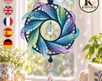 Puede incluir: Un adorno de pared circular de ganchillo con un anillo azul oscuro adornado con hojas radiantes de colores en tonos azul, verde y morado. Una cuenta de cristal facetado cuelga en el centro, con un cristal en forma de lágrima debajo.