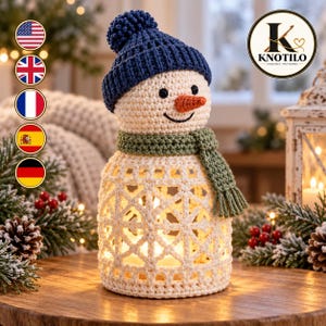 May include: A crocheted snowman lantern featuring a cream-colored body with an openwork snowflake pattern, a dark blue beanie, a sage green scarf, and an orange carrot nose. The logo KNOTILO CROCHET PATTERNS is visible in the upper right corner.