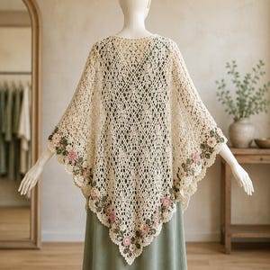 May include: A cream-colored crochet shawl featuring an intricate diamond-patterned openwork design. The edges are adorned with a decorative border of small, three-dimensional pink flowers and green leaves, draped over a solid olive green dress.