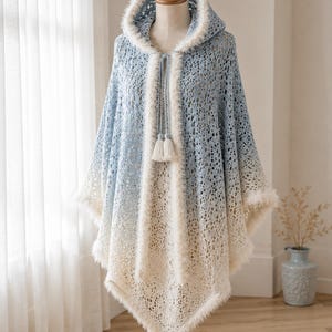 Könnte beinhalten: Ein gehäkelter Poncho mit Kapuze und einem Farbverlauf von Hellblau oben bis Cremeweiß unten. Das Kleidungsstück ist mit einem flauschigen weißen Rand an der Kapuze und den Kanten versehen und verfügt über einen Kordelzug mit zwei cremefarbenen Quasten.