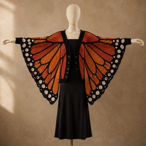 May include: A crochet cardigan featuring a monarch butterfly wing pattern in vibrant orange, black, and white. The garment has long sleeves, a black trim, and a front button closure, displayed on a mannequin wearing a plain black dress.