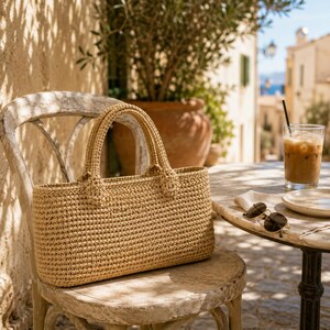 Puede incluir: Un bolso tote rectangular de color beige, tejido a mano al crochet, con dos asas resistentes a juego. El bolso presenta un patrón de puntos apretados y uniformes en toda su estructura, lo que le otorga una forma firme. La superficie es texturizada.