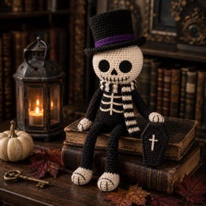May include: A crocheted skeleton doll wearing a black top hat with a purple band and a black and white striped scarf. The doll has a cream-coloured skull with black eye sockets and a stitched mouth, holding a small black coffin with a white cross.