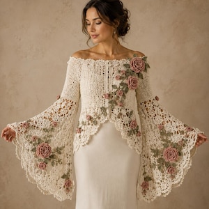 May include: An off-the-shoulder cream crochet cardigan featuring long, flared bell sleeves and an intricate openwork pattern. The garment is adorned with three-dimensional dusty pink crocheted roses and green leaves trailing across the front and sleeves.