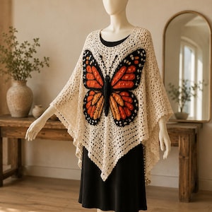 May include: A cream-colored crochet poncho featuring a large, detailed monarch butterfly appliqué on the front. The butterfly displays vibrant orange, yellow, and black thread work. The poncho has an open-knit texture and a pointed hemline.