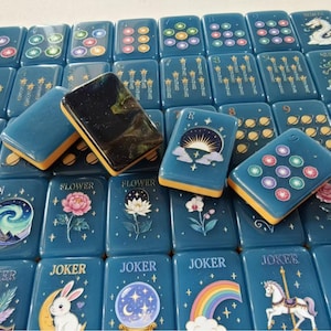 May include: A collection of rectangular mahjong tiles with a deep blue base and yellow edges. The tiles feature various celestial, floral, and whimsical illustrations, including a rabbit, a carousel horse, a rainbow, and a dark, glittery tile.