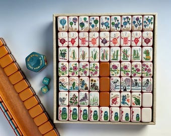 Puede incluir: Un juego de mahjong con fichas blancas que presentan ilustraciones coloridas de globos aerostáticos, linternas, bambú, flores y figuras. El set incluye un soporte de madera, dos dados verdes pequeños, un recipiente hexagonal y fichas naranjas.