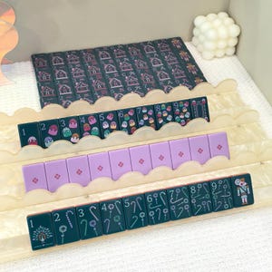 May include: A set of rectangular mahjong tiles arranged on a tiered, scalloped-edge acrylic rack. The tiles feature various designs including numbers, colorful sweets, floral patterns, candy canes, and a nutcracker figure in dark green and purple.
