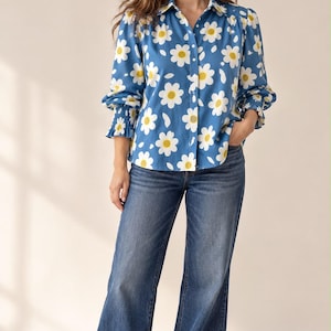 Puede incluir: Una camisa de manga larga con botones en color azul que presenta un estampado floral de margaritas blancas y amarillas, combinada con pantalones vaqueros azules de pierna ancha y zapatillas blancas. La camisa tiene cuello clásico y puños elásticos.