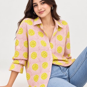 Puede incluir: Una camisa de manga larga con botones que presenta un patrón repetitivo de rodajas de limón amarillo brillante sobre un fondo rosa claro con finas rayas blancas horizontales y verticales que forman un diseño de cuadrícula.