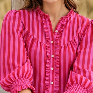 May include: A button-down blouse featuring vertical stripes in alternating shades of hot pink and magenta. The garment includes a ruffled collar, a ruffled placket along the front buttons, and three-quarter length sleeves with smocked cuffs.