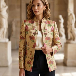 May include: A gold-toned blazer featuring a vibrant floral brocade pattern in shades of pink, red, and green. The jacket has a structured mandarin collar, long sleeves, and an open front design, worn over a simple white camisole and black trousers.