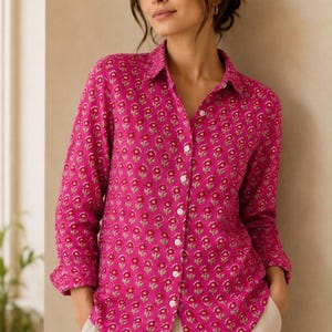May include: A long-sleeved button-down shirt in a vibrant magenta pink colour featuring a repeating pattern of small, stylised red and white flowers with green stems. The shirt has a classic collar, a front button placket, and a curved hemline.