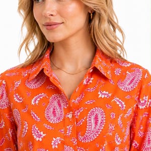 Pode incluir: Uma camisa de manga comprida com botões numa cor laranja vibrante com um padrão paisley branco e rosa repetitivo. A camisa tem um colarinho clássico, uma carcela frontal com botões brancos e um corte estruturado nos ombros.