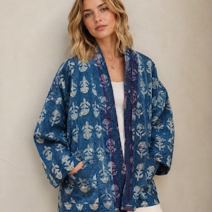 May include: A blue quilted jacket featuring a repeating light-coloured floral block print pattern. The jacket has an open front design with a dark blue trim along the collar and two front pockets. The fabric has a textured, stitched appearance.