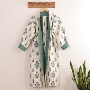 May include: A long-sleeved, knee-length quilted robe in off-white featuring a repeating pattern of blue floral motifs and green leaves. The garment includes a solid sage green inner lining, a shawl collar, and two front patch pockets on the exterior.