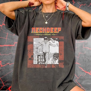 Puede incluir: Una camiseta gris carbón de manga corta con un estampado gráfico que incluye el texto "NECK DEEP" y "THE PEACE AND PANIC TOUR". El diseño muestra a una persona con traje frente a un horizonte urbano y una gran nube, en blanco y negro.
