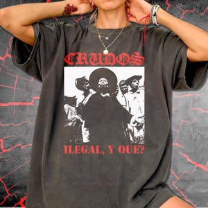 Peut inclure: Un t-shirt gris anthracite à manches courtes avec une impression graphique centrale en noir et blanc représentant un groupe de personnes. Au-dessus du graphique se trouve le texte rouge CRUDOS et en dessous le texte rouge ILEGAL, Y QUE?