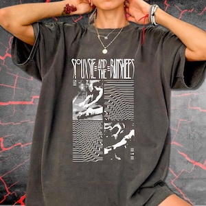 May include: A charcoal gray short-sleeve t-shirt featuring a white graphic print. The design includes the text Siouxsie and the Banshees above four square panels containing abstract wave patterns, textured lines, and high-contrast silhouettes.