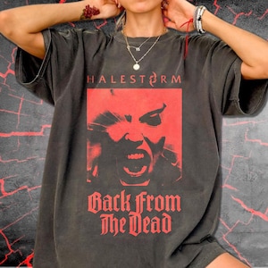 May include: A charcoal gray short-sleeve t-shirt featuring a red graphic print. The design includes the text HALESTORM above a stylized face with an open mouth, and the text Back from The Dead printed in a bold red font below the graphic.