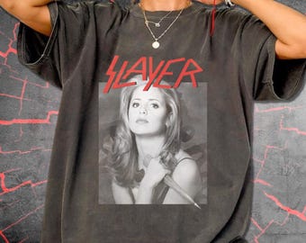 May include: A charcoal gray short-sleeve t-shirt featuring a black and white photographic print of a woman holding a stake. Above the image, the word SLAYER is printed in a bold red font. The shirt has a crew neckline and a relaxed fit.