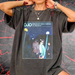 Puede incluir: Una camiseta gris carbón de manga corta con un estampado gráfico rectangular. El estampado muestra el texto DJO BACK ON YOU WORLD TOUR 2025 WITH POST ANIMAL sobre una escena nocturna de tres personas bajo farolas.