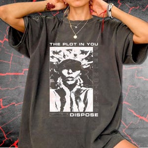 May include: A charcoal grey short-sleeve t-shirt featuring a white graphic print of a person with a crown-like headpiece. The text reads THE PLOT IN YOU above the image and FEEL NOTHING DISPOSE below, with vertical text along the right side.