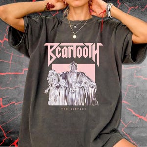 Puede incluir: Una camiseta de manga corta gris carbón con un estampado gráfico rosa claro. El diseño muestra el texto Beartooth en una fuente estilizada sobre una ilustración de una figura sentada en un trono, con el texto THE SURFACE impreso debajo.