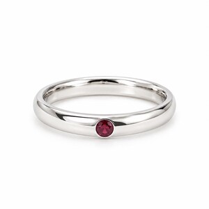 Può includere: Un anello a fascia in metallo color argento lucido con una singola piccola pietra preziosa rotonda di colore rosso intenso, incastonata a filo al centro della fascia. L'anello ha un profilo liscio e arrotondato con una finitura brillante.
