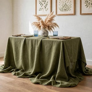 May include: A rectangular olive green tablecloth draped over a table, featuring a textured fabric surface. On the table, there is a central vase filled with dried pampas grass, two blue drinking glasses, two white plates, and two folded napkins.