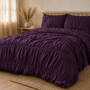 Puede incluir: Un juego de cama de color morado intenso que incluye un edredón y dos fundas de almohada a juego. La tela presenta una textura fruncida con detalles de volantes horizontales en la superficie del edredón y en el frente de las fundas.