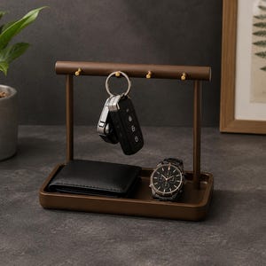 May include: A dark brown metal valet tray featuring a horizontal bar with four small gold-toned hooks. The tray holds a black leather wallet and a silver-toned chronograph wristwatch, while a set of car keys hangs from one of the gold hooks.