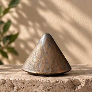 May include: A conical object with a textured surface featuring a blend of metallic bronze and dark gray tones. The finish appears weathered and uneven, with a smooth, rounded peak and a wide, circular base that tapers toward the top point.