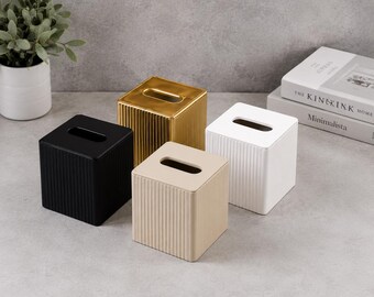 Könnte beinhalten: Vier quadratische Taschentuchboxen mit vertikal gerippter Textur. Das Set enthält eine schwarze, eine goldfarbene, eine beige und eine weiße Box, die jeweils eine rechteckige Öffnung an der Oberseite zur Entnahme der Taschentücher aufweisen.