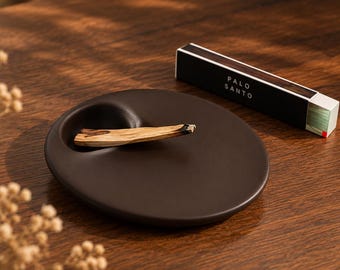 Könnte beinhalten: Ein dunkelbrauner, kreisförmiger Räucherstäbchenhalter aus Keramik mit einer Vertiefung, in der ein helles Stück Palo Santo-Holz liegt. Daneben befindet sich eine rechteckige schwarze Schachtel mit dem weißen Text Palo Santo.